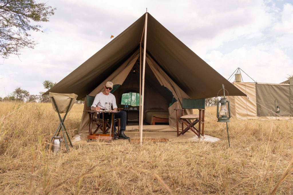 Tansania Zeltsafari – Ihr privates Camp in der Serengeti Reisender sitzt schreibend vor Zelt, Blick aufs Bett, Select-Zelt-Safari, Serengeti Tansania, Globetrotter Select